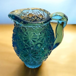 May include: A teal-coloured glass creamer with a starburst pattern. The creamer has a handle and a spout, and the glass has a textured surface. It is likely from the mid-20th century and is a decorative item.