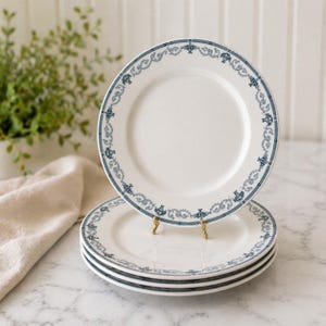 Pode incluir: Uma pilha de quatro pratos de cerâmica branca com uma borda decorativa azul. A borda apresenta um padrão repetitivo de arabescos ornamentados e pequenos motivos de coroa. Os pratos estão empilhados em uma superfície de mármore.