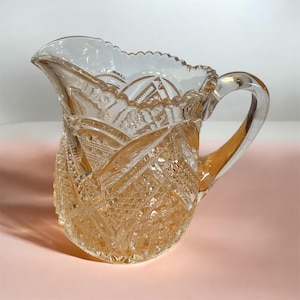 May include: A clear, antique glass pitcher with a decorative, cut-glass pattern. The pitcher has a scalloped rim and a curved handle. The glass has a slight amber tint, and the pitcher is set against a pink background. The pitcher is likely used for serving beverages.