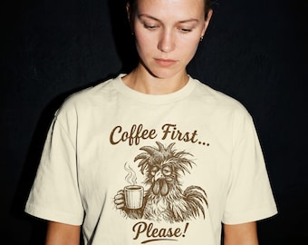 May include: A cream-coloured t-shirt with a brown illustration of a rooster holding a coffee cup, and the text "Coffee First... Please!". The shirt has a crew neck and short sleeves. The design is a humorous take on morning routines.