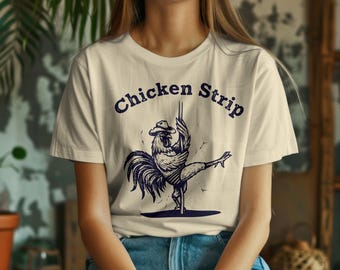 May include: Cream-coloured t-shirt with a navy blue graphic of a rooster wearing a cowboy hat, pole dancing. The text "Chicken Strip" is arched above the rooster. The shirt is paired with blue jeans.