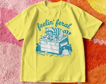 May include: Yellow t-shirt featuring a teal graphic of a raccoon in a dumpster, with the text "feelin' feral" above. The design includes trash bags and fish bones. The shirt is made of a soft material. The design is printed in teal.