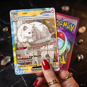 Può includere: Una carta Pokémon Luna con un'illustrazione di un soffice gatto bianco. La carta presenta il testo "Velvet Paw" e "Fur Storm". È tenuta accanto a una carta Pokémon rosa con un design Pokéball.