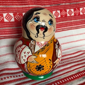 Può includere: Una matrioska in legno raffigurante un personaggio dipinto con baffi neri, occhi azzurri e capelli scuri. La figura indossa una camicia bianca con motivi floreali rossi e un gilet marrone, mentre tiene in mano un grande strumento a corda arancione.