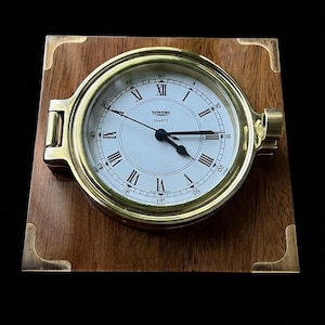 May include: A round quartz clock with a brass frame and white face featuring black Roman numerals and the text Talamec Quartz. The clock is mounted on a square wooden plaque with brass corner accents and a side hinge mechanism for opening.