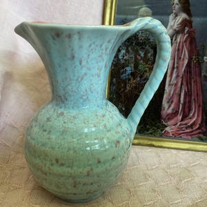 May include: A light blue and green ceramic jug with a glossy crackle glaze finish and speckled brown accents. The vessel features a wide, flared rim, a rounded body, and a textured, twisted handle extending from the side to the neck.