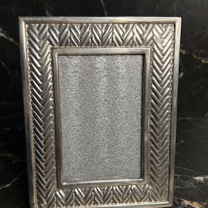 May include: A rectangular silver-toned picture frame featuring a prominent embossed herringbone pattern along its border. The center of the frame contains a textured, shimmering silver insert, providing a metallic finish to the overall design.