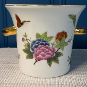 May include: A white porcelain vessel with a flared rim and two gold-toned side handles. The exterior features a colorful floral arrangement of pink, blue, and orange blossoms with green leaves, accented by two small butterflies in orange and green.