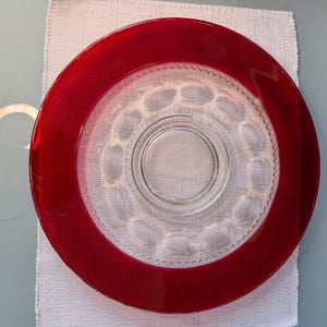 Puede incluir: Un plato de vidrio transparente con un borde rojo. El plato tiene un diseño circular en el centro y está colocado sobre un paño blanco. El plato probablemente se utiliza para servir alimentos.