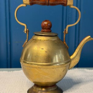 May include: A brass kettle with a spherical body, a pedestal base, and a curved spout. The kettle features a hinged handle with a dark brown wooden grip and gold-toned metal accents, topped with a matching dark brown wooden knob on the lid.