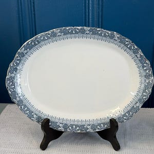 May include: An oval ceramic serving platter featuring a white center and a wide border decorated with a blue and white floral pattern. The rim has a scalloped edge, and the platter is displayed on a black stand, showcasing its detailed design.