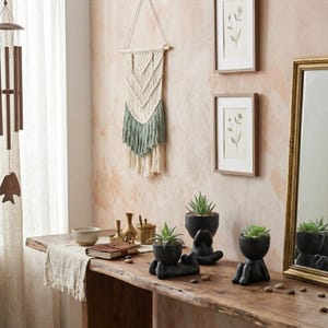 May include: Two matte black ceramic planters shaped like figures sitting with legs extended, each holding a green succulent plant. The planters are placed on a wooden surface alongside a small bowl, a closed notebook, and a brass-colored container.