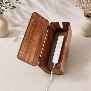 May include: A rectangular wooden cable management box with a hinged lid, featuring a dark brown wood grain finish. Inside the open box sits a white power strip with multiple outlets. A white power cord extends from the bottom of the wooden container.