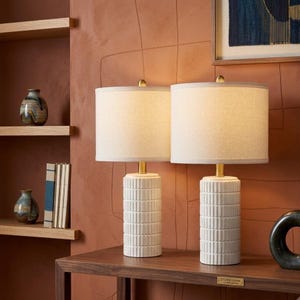 May include: Two table lamps with cylindrical white ceramic bases featuring a vertical ribbed texture. Each lamp has a brass-colored metal neck and a cylindrical off-white fabric drum shade. The lamps are positioned on a wooden surface.