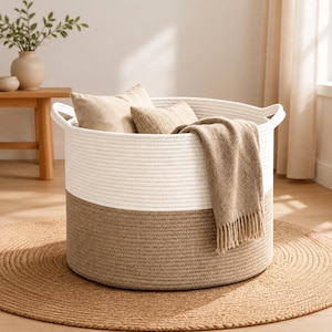 May include: A large, round storage basket crafted from coiled rope, featuring a two-tone design with a white upper section and a tan lower section. The basket includes two integrated side handles and contains two beige pillows and a fringed throw blanket.