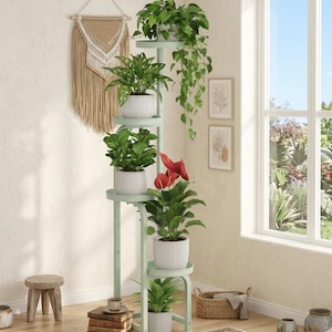 May include: A light green metal plant stand with six circular tiers arranged in a vertical, staggered structure. The stand holds various potted plants, including a trailing vine, a red anthurium, and several green leafy plants in white ribbed pots.