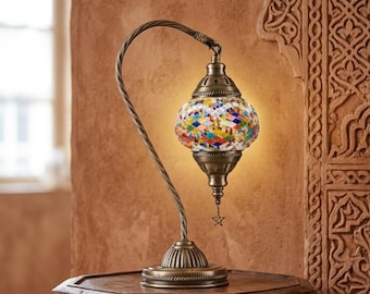 May include: A table lamp with a curved, twisted bronze-toned metal stand and a base. The lamp features a globe-shaped shade made of multi-colored glass mosaic tiles in a diamond pattern, with a small metal star charm hanging from the bottom.