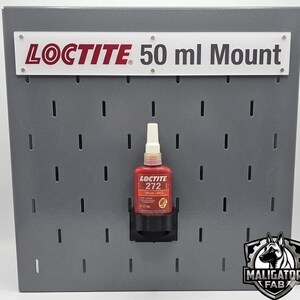 Puede incluir: Un panel perforado de metal gris con un soporte negro que contiene una botella roja de Loctite 272. El letrero de arriba dice "LOCTITE 50 ml Mount". El panel perforado tiene múltiples ranuras para el montaje. Una esquina inferior derecha tiene un logotipo.