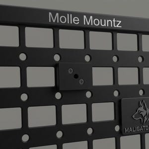 Könnte beinhalten: Schwarzes Molle Mountz-Panel mit einem Raster aus rechteckigen und kreisförmigen Öffnungen. Eine kleinere schwarze Platte ist mit Schrauben befestigt. Der Text "Molle Mountz" steht oben. Ein Logo mit einem Wolfskopf und dem Text "MALIGATOR FAB" befindet sich unten rechts.