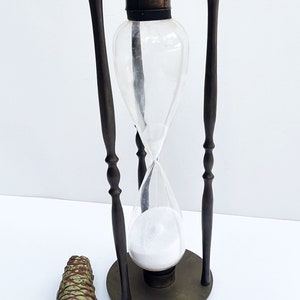 May include: A brass hourglass with a clear glass bulb and white sand. The hourglass is on a stand with two black wooden posts.