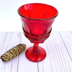 May include: A red glass goblet with a faceted design. The goblet is sitting on a white wooden surface.