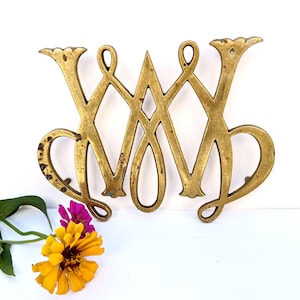 May include: A gold-toned metal trivet with an intricate design. The trivet is shaped like the letter 'W' and has a decorative, ornate style.