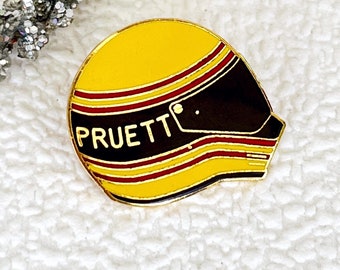 May include: A yellow and black racing helmet pin with the name "Pruett" in gold lettering.