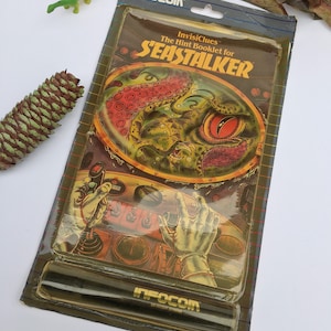 May include: A brown and green game booklet with the title "InvisiClues: The Hint Booklet for Seastalker" and a picture of a green and red octopus. The booklet is packaged in a clear plastic sleeve with a black and white label that says "Infocom".