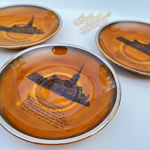 May include: Three brown and gold ceramic saucers with a silver rim. Each saucer features a black and white image of Salisbury Cathedral with a quote about the cathedral.