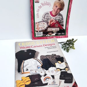 May include: Two cross-stitch pattern books. The top book is red and features a woman cross-stitching a holiday-themed design. The bottom book is beige and features a variety of cross-stitch designs on different items, including a t-shirt, a pair of slippers, and a tote bag. The text on the bottom book reads "Cross Stitch Waste Canvas Designs".