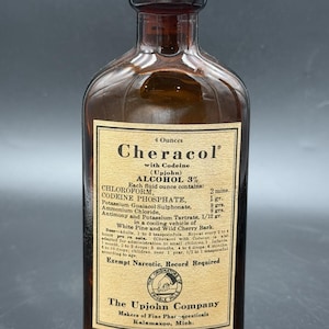 May include: A vintage amber glass bottle with a cork stopper, featuring a beige paper label for Cheracol with Codeine. The label lists ingredients including chloroform, codeine phosphate, and alcohol, with The Upjohn Company branding.