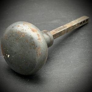 May include: A round, dark gray metal door knob with visible rust spots and a weathered texture. The knob features a long, square-shaped metal spindle extending from the back, which includes a series of small holes along its length for installation.