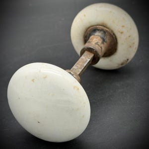 May include: A vintage door knob set featuring two round, white porcelain handles connected by a square metal spindle. The white surfaces show signs of age with small brown spots and discoloration, while the metal components exhibit a dark, weathered patina.