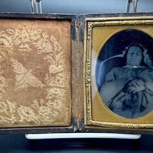 May include: An antique daguerreotype photograph in a hinged case. The left side features a tan velvet lining with an embossed floral wreath and butterfly. The right side contains a portrait of a person with eyes closed, framed in an oval gold mat.