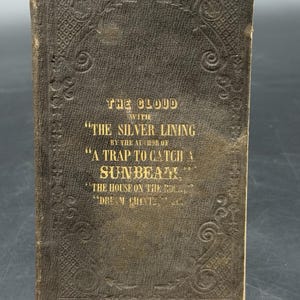 May include: A dark brown antique book with an embossed decorative border on the cover. The gold text reads: The Cloud with The Silver Lining by the author of A Trap to Catch a Sunbeam, The House on the Rock, Dream Chintz, etc.