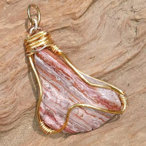 May include: A teardrop-shaped pendant featuring a polished, banded stone with alternating layers of reddish-brown and white. The stone is secured in a gold-colored wire wrap that forms a decorative frame and includes a small circular bail at the top.