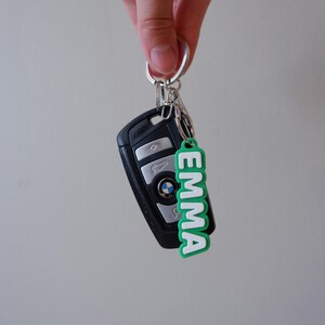 Op de afbeelding: Een zwarte autosleutel met een zilveren sleutelring en een groen-witte sleutelhanger met de tekst "EMMA". De sleutelhanger heeft het BMW-logo.