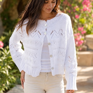 May include: A white long-sleeve knit cardigan featuring a round neckline, a button-down front with five circular buttons, and intricate openwork patterns across the chest and sleeves, finished with ribbed cuffs and a ribbed hemline.