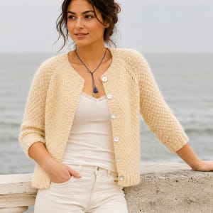 May include: A light beige, textured knit cardigan with three-quarter sleeves and button closures. The cardigan is worn over a white tank top and paired with white pants. A blue necklace adds a pop of color. The overall style is casual and comfortable.