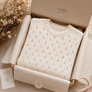 May include: Cream-colored sweater with a diamond pattern, presented in a box. The sweater has a crew neck and is folded neatly inside the box, which is lined with tissue paper. The box has the text "LUNE ATELIER" on the lid.