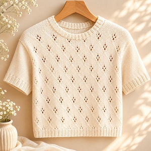 May include: Cream-colored short-sleeve knit top with a diamond-shaped openwork pattern. The top has a crew neck and is displayed on a wooden hanger. The knitwear is a soft, neutral color, suitable for various outfits.