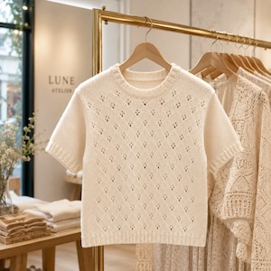 May include: Cream-colored short-sleeved sweater with a diamond-shaped openwork pattern. The sweater is hanging on a wooden hanger. The word "LUNE ATELIER" is visible in the background.