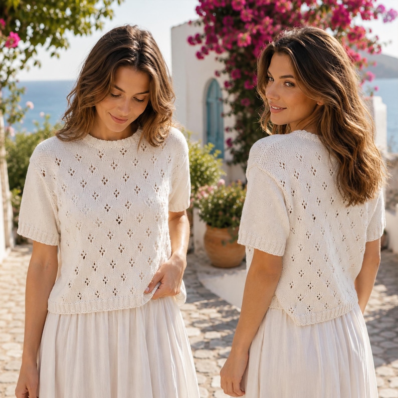 May include: Two women wearing matching cream-colored knit tops with diamond-shaped cutouts and short sleeves, paired with long cream skirts. The background features a coastal scene with a white building, blue water, and pink flowers.