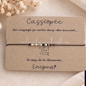 Peut inclure: Un bracelet en cordon noir fin avec une séquence de petites perles blanches, noires et métalliques en code Morse, fixé sur une carte de présentation en papier kraft rectangulaire portant le texte Cassiopée et Enigma imprimé.