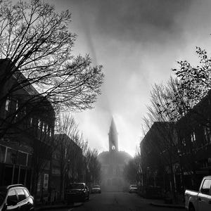 May include: Black and white photograph of a street scene with buildings and bare trees lining the road. A church steeple is visible in the distance, shrouded in fog. Cars are parked along the street.