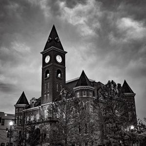 May include: Black and white photograph of a historic brick building with a clock tower. The building has multiple turrets and arched windows. The sky is overcast, and trees line the street in front of the building.