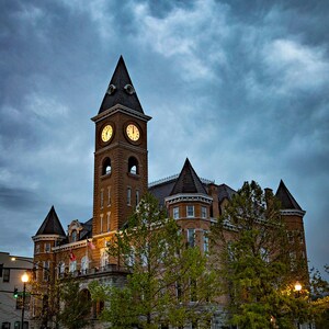 May include: A stately brick building with a clock tower under a cloudy sky. The tower has two illuminated clock faces. The building has multiple turrets and is surrounded by trees and a grassy area. The image is taken at dusk.