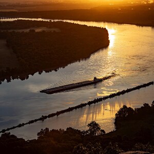 May include: An aerial view of a long, narrow barge traveling down a wide river during a golden sunset. The water reflects the warm orange light of the sun, while dark silhouettes of trees line the riverbanks and a stone jetty extends into the water.