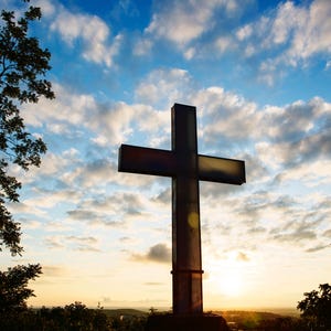 Op de afbeelding: Een groot, donker kruis tegen een levendige lucht vol wolken. Het kruis staat hoog, met de ondergaande zon op de achtergrond, wat een dramatische en spirituele scène creëert. Bomen omlijsten de zijkanten van de afbeelding.