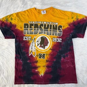May include: A short-sleeved t-shirt featuring a tie-dye pattern in yellow, burgundy, and black. The front displays the text WASHINGTON REDSKINS EST. 1932, a circular logo with a profile of a person wearing a feather headdress, and a letter N.